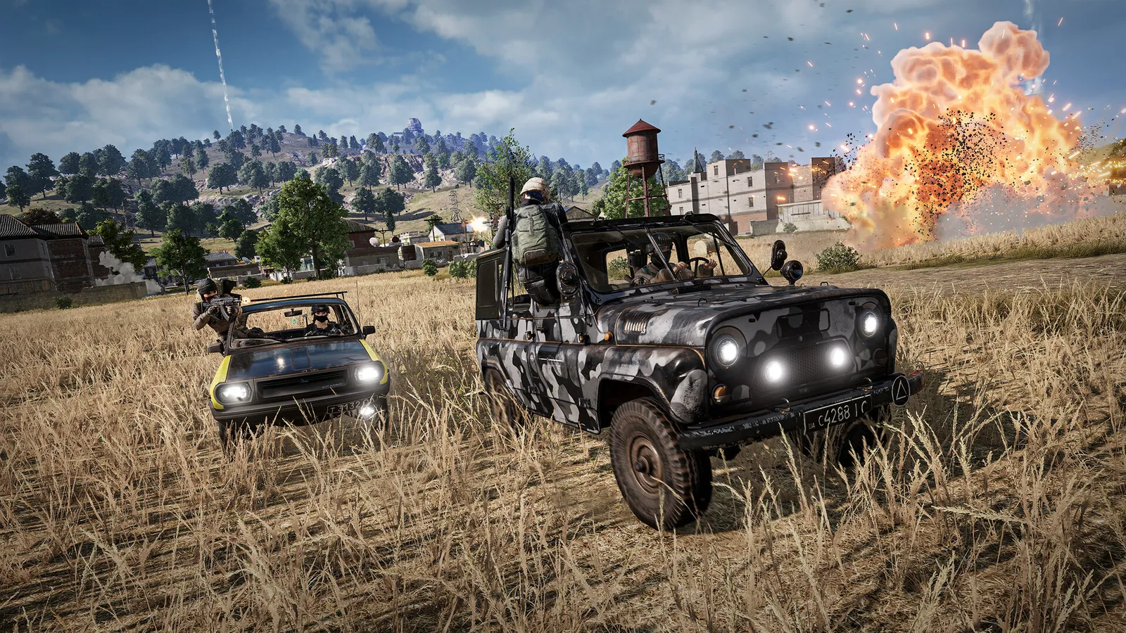 Veículos com jogadores armados atravessam um campo seco, com uma explosão ao fundo em PUBG: BATTLEGROUNDS.